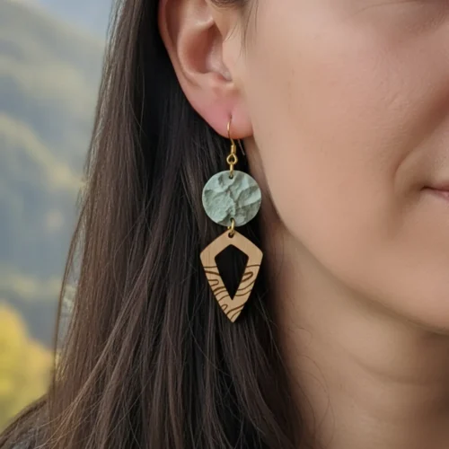 Boucles d'oreilles Peaks en bois avec pendentif gris clair vert au relief de montagnes imprimées en 3D dans du plastique recyclé et forme pointue en bois suisse, portées par femme avec cheveux longs noirs et ciel flou en arrière-plan